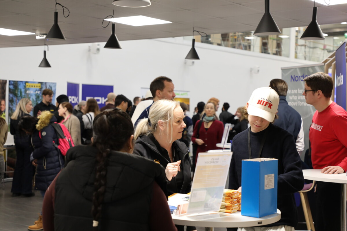 Ihmiset ovat vuorovaikutuksessa kiireisessä sisätiloissa järjestettävässä tapahtumassa, jossa on kojuja ja banderolleja. Kaksi ihmistä keskustelee pöydän ääressä, jossa on esitteitä ja välipaloja, kun muut kävelevät ja puhuvat taustalla. Yläpuolella roikkuvat kattovalaisimet.