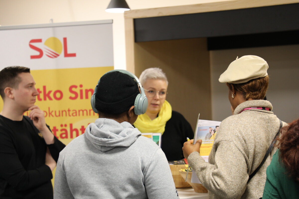 Joukko ihmisiä seisoo ja puhuu sisätiloissa järjestettävässä tapahtumassa. Lyhyet harmaat hiukset ja silmälasit päässä oleva nainen puhuu pöydän takana, kun muut kuuntelevat ja katselevat esitteitä. Taustalla näkyy SOLin banneri.