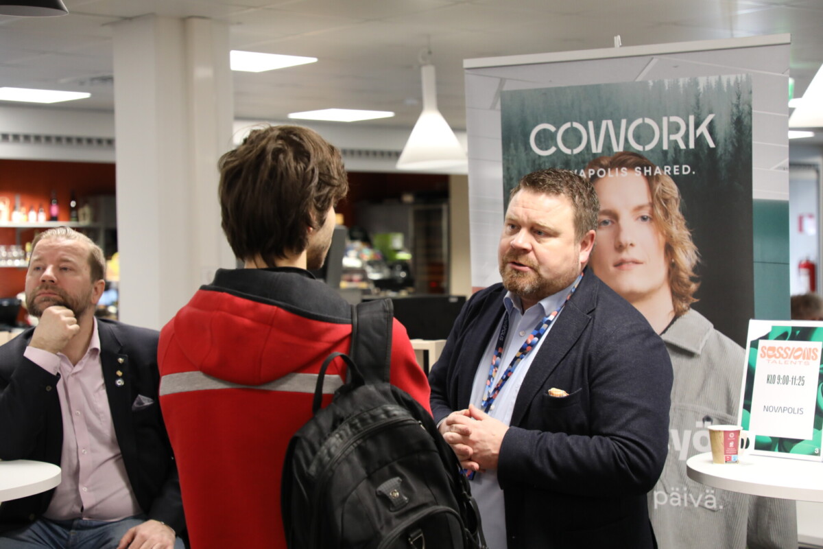 Kaksi miestä keskustelee verkostoitumistilaisuudessa korkean "COWORK"-banderollin vieressä. Toisella miehellä on reppu kädessään, kun taas toinen pukumies kuuntelee tarkkaavaisesti. Toinen henkilö istuu pöydän ääressä taustalla.
