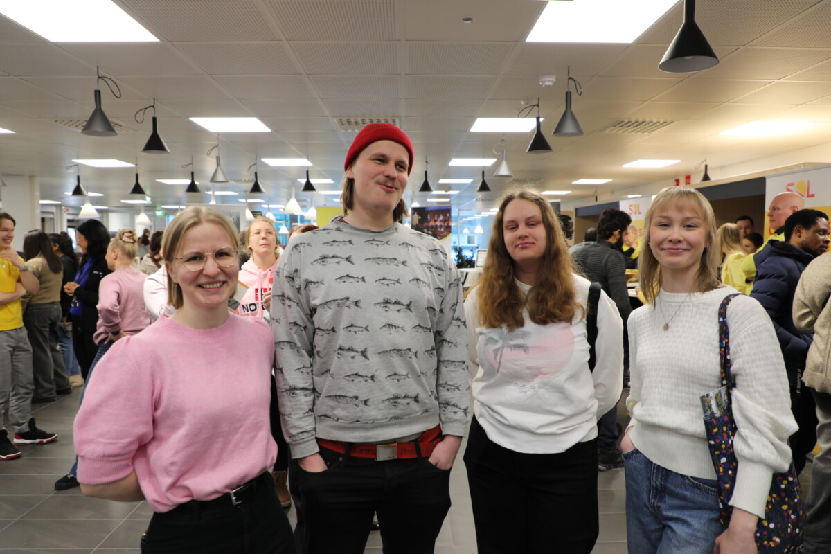 Neljä ihmistä seisoo yhdessä sisätiloissa hymyillen kameralle. Taustalla on lisää ihmisiä, ja ympäristö vaikuttaa vilkkaalta sosiaaliselta tapahtumalta tai kokoontumiselta, mahdollisesti kahvilassa tai yhteisessä tilassa.