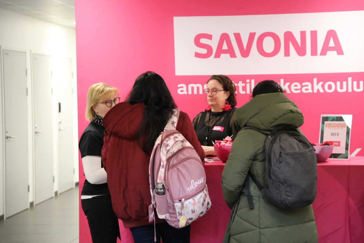Kolme reppupäistä ihmistä puhuu kahdelle naiselle kirkkaan vaaleanpunaisen Savonia-infotiskin käytävällä. Tiskillä olevilla naisilla on nimilaput, ja taustalla on Savonia-logo vaaleanpunaisella seinällä.