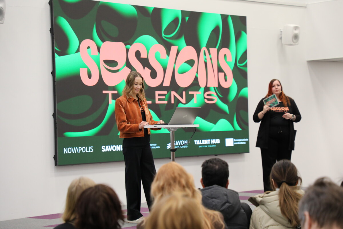 Nainen puhuu korokkeella yleisön edessä, ja hänen takanaan on värikäs "Sessions Talents" -kyltti. Toinen nainen seisoo vieressä ja tulkkaa viittomakielelle.