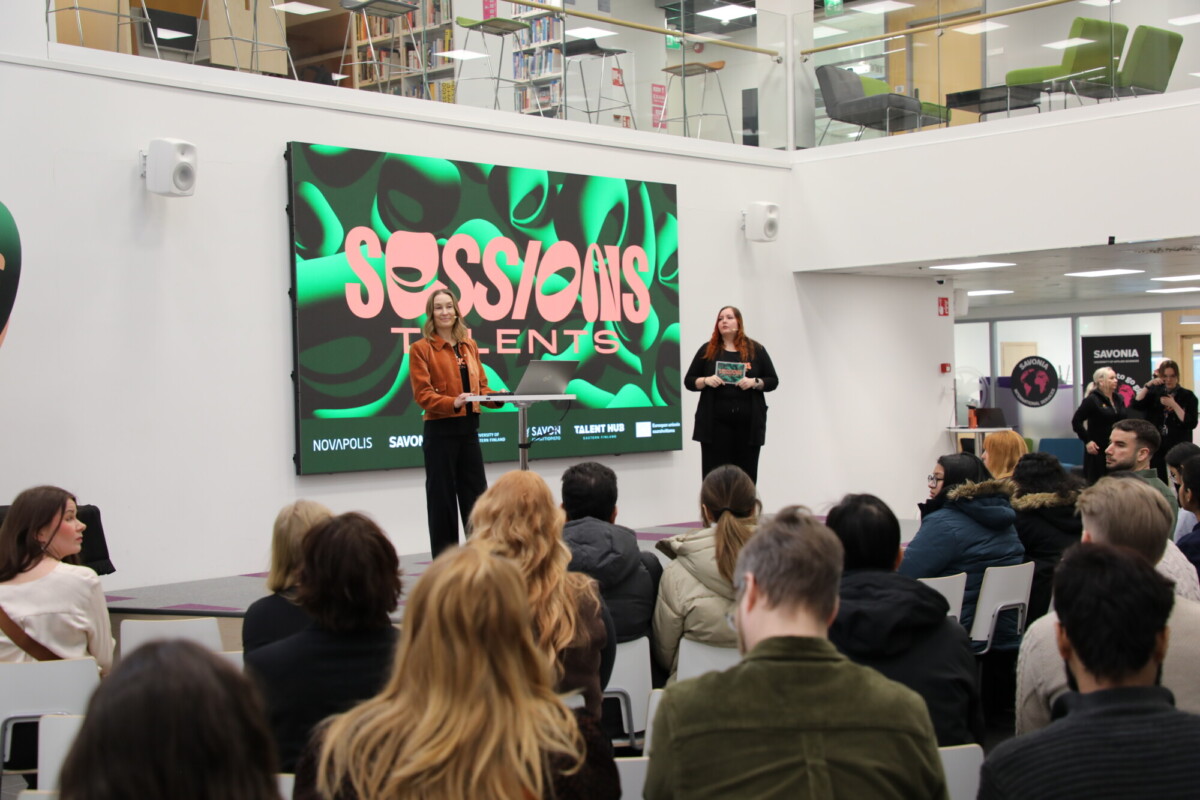 Kaksi naista seisoo lavalla yleisön edessä modernissa sisätilassa, ja heidän takanaan on suuri näyttö, jossa lukee lihavoidulla, värikkäällä tekstillä Sessions Talents.