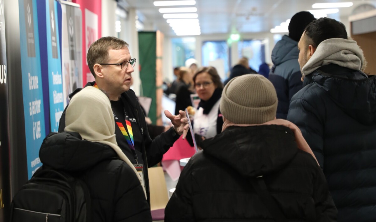Silmälasipäinen mies puhuu pienelle joukolle talvivaatteisiin pukeutuneita ihmisiä sisätiloissa järjestettävässä tapahtumassa, jonka taustalla on kojuja ja tiedotusbanderolleja. Muita osallistujia näkyy vilkkaalla käytävällä.