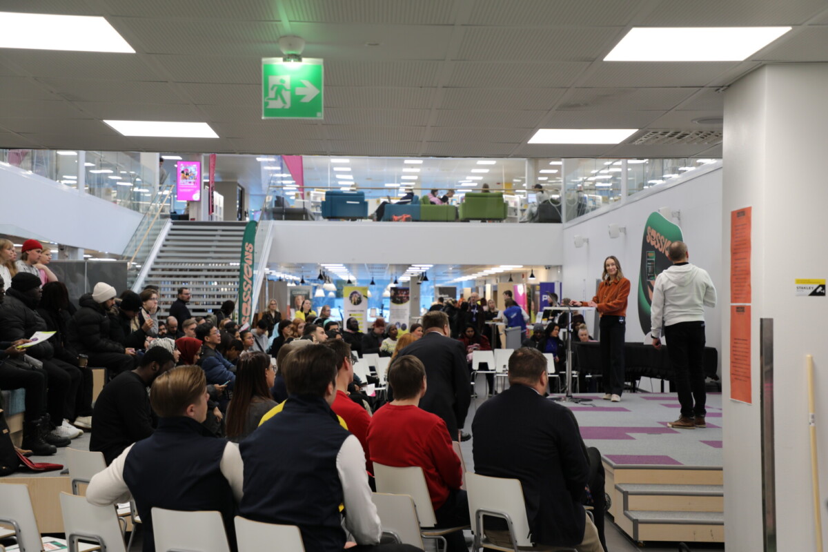 Yleisö istuu modernissa sisätilassa, vastapäätä lavaa, jossa nainen puhuu mikrofoniin ja mies seisoo vieressä. Yleisö seuraa tarkkaavaisesti; taustalla näkyy portaita ja oleskelutiloja.