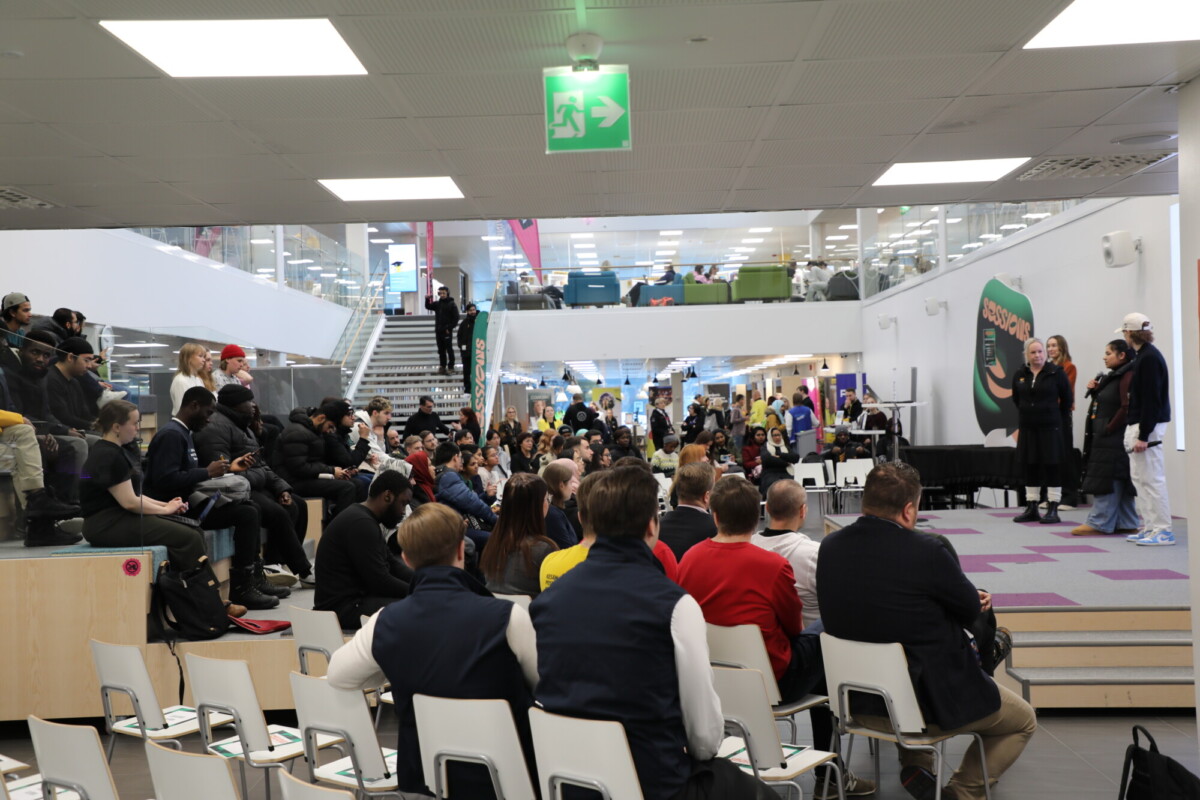 Suuri joukko ihmisiä istuu ja kuuntelee puhujia lavalla modernissa, avoimessa sisätilassa, jossa on portaat, lasiseinät ja kirkas valaistus. Etualalla on tyhjiä tuoleja.