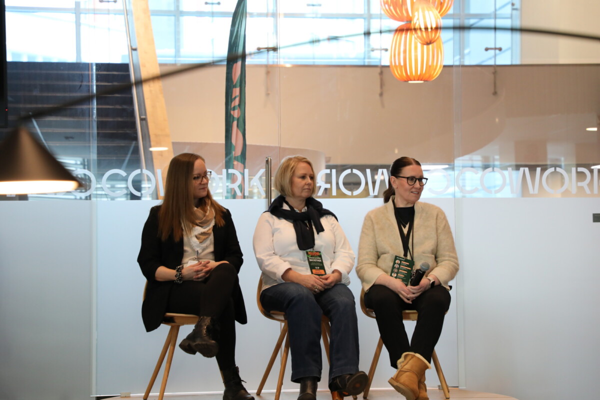 Kolme naista istuu tuoleilla modernissa sisätilassa, jossa on lasiseinät. He näyttävät osallistuvan paneelikeskusteluun. Taustalla on COWORK-teksti ja roikkuvia valoja.