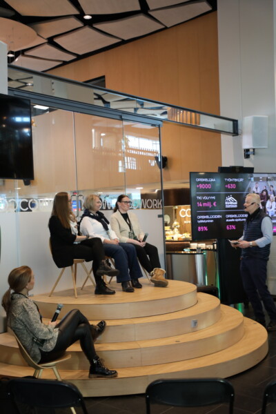 Kolmen naisen muodostama paneeli istuu puisella lavalla, kun toinen nainen istuu mikrofonin kanssa lavan ulkopuolella. Mies seisoo kahden suuren näyttöruudun vieressä, joissa näkyy tilastoja ja tekstiä, ja keskustelee paneelin kanssa modernissa sisätilassa.