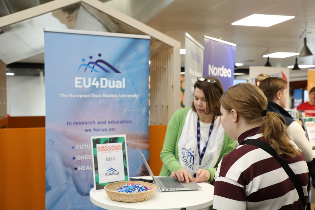 Kaksi naista seisoo kojulla, jonka takana on EU4Dual-banderolli. Toinen käyttää kannettavaa tietokonetta ja toinen katsoo vierestä. Pöydällä on kulhollinen karkkia ja tiedotuskylttejä kiireisessä sisätiloissa järjestettävässä tapahtumassa.