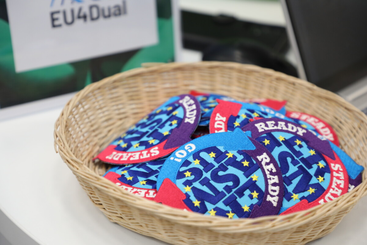 Pajukori, joka on täynnä värikkäitä kirjailtuja laastareita, joissa lukee "READY, STEADY, GO", on asetettu pöydälle "EU4Dual" -kyltin viereen.