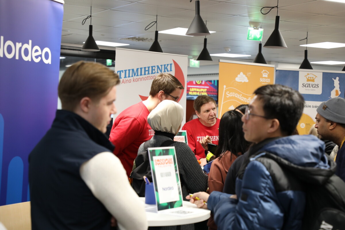 Ihmiset kerääntyvät sisäilmatapahtuman infopisteiden ympärille. Useita banderolleja ja kylttejä on näkyvissä, ja ihmiset keskustelevat. Yhdellä henkilöllä on kädessään esite, ja tapahtuman henkilökunnalla on punaiset paidat.