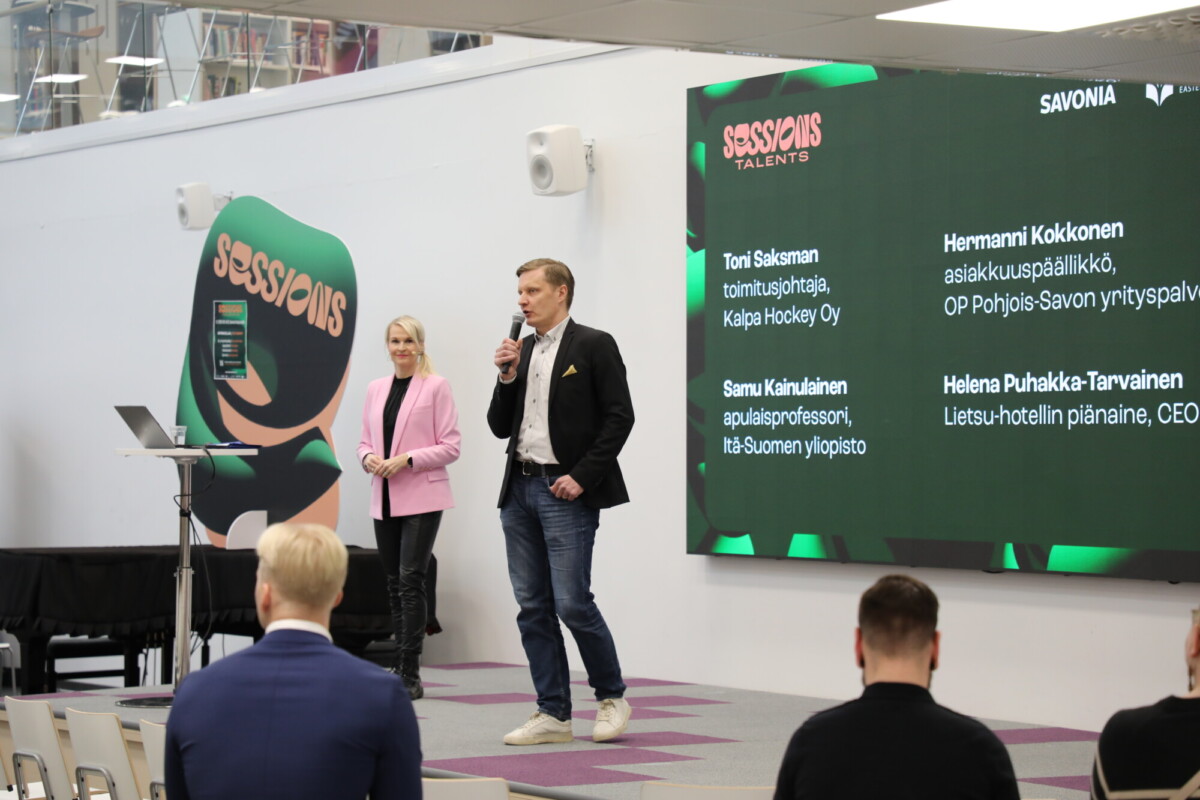 Mies puhuu mikrofoniin lavalla naisen vieressä, jolla on leikepöytä kädessään. Heidän takanaan suurella näytöllä näkyvät nimet ja tittelit Sessions Talents -tapahtumassa. Yleisössä istuu muutama ihminen.