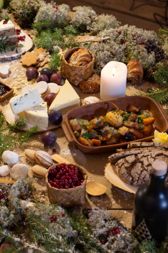 Maalaismainen pöytä, jossa on erilaisia juustoja, leipää, paahdettuja vihanneksia, macaroneja, marjoja, keksejä ja sytytetty valkoinen kynttilä, jota ympäröivät vehreät ja sammaloituneet koristeet.