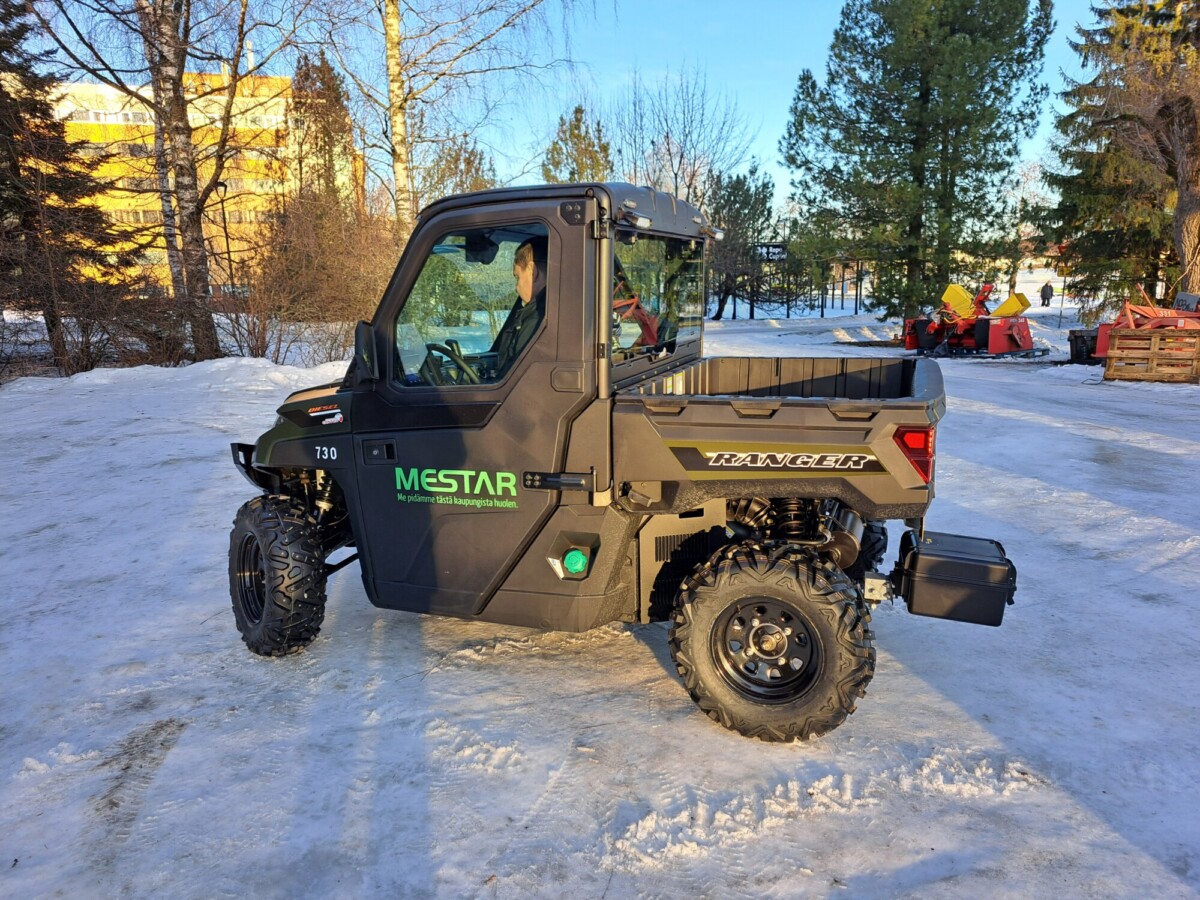 Musta Mestar Ranger -hyötyajoneuvo on pysäköity lumiselle maalle, ja taustalla näkyy puita ja rakennuksia aurinkoisena päivänä.