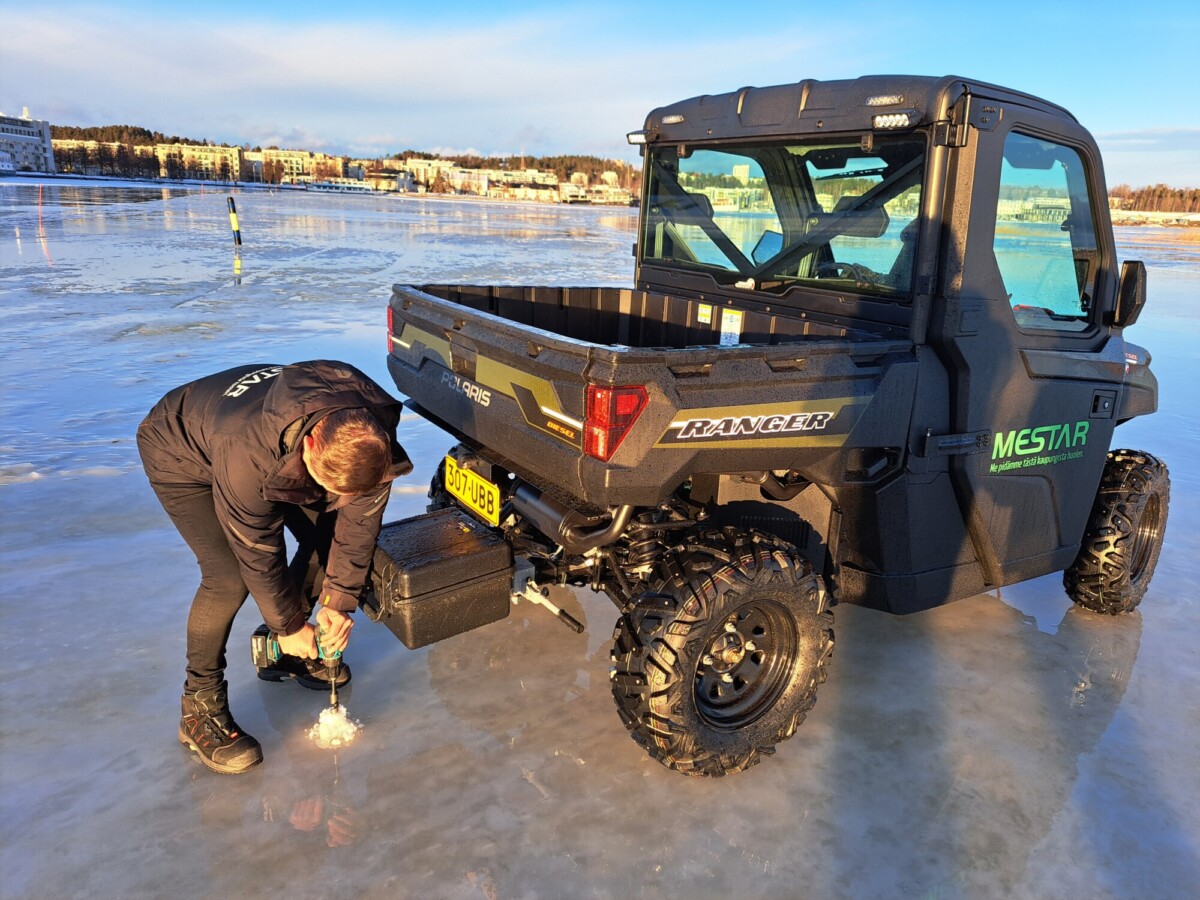 Henkilö poraa reikää paksuun jäähän sähkötyökalulla mustan hyötyajoneuvon vieressä, jossa on merkinnät Ranger ja Mestar. Ajoneuvo on pysäköity jäiselle pinnalle lähelle kaupunkia, jonka taustalla on rakennuksia.