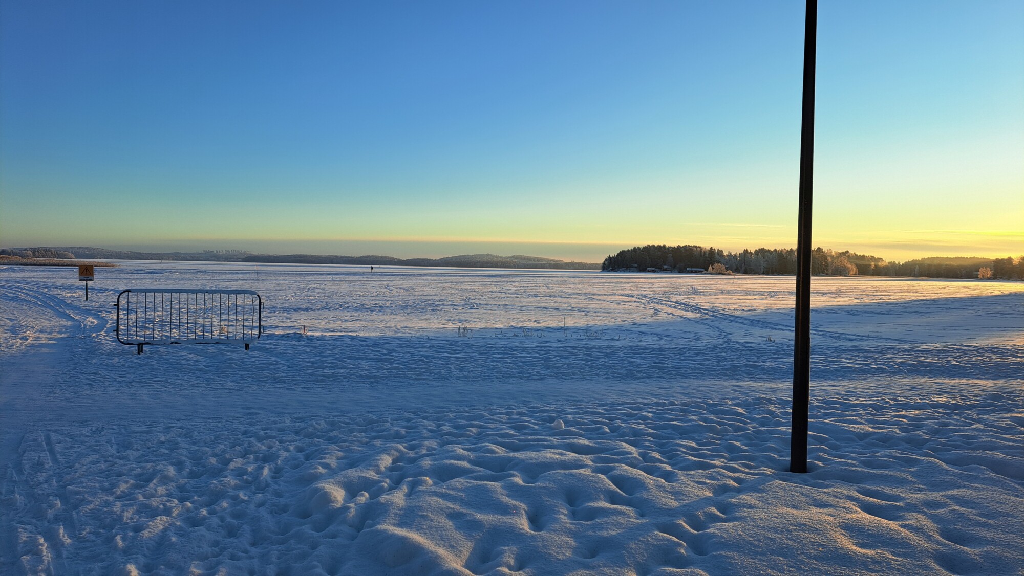 Luminen pelto kirkkaan sinisen taivaan alla auringonnousun tai -laskun aikaan, etualalla jalanjäljet lumessa, metallieste, roskakori ja yksittäinen musta pylväs. Kaukana näkyy puita ja kukkuloita.