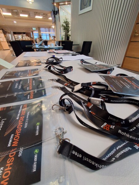 Pöytä tapahtuman ilmoittautumisalueella, jossa on kaulaliinat, nimikyltit ja tiedotuslehtiset siististi järjestettyinä. Taustalla näkyy moderni rakennuksen sisustus.