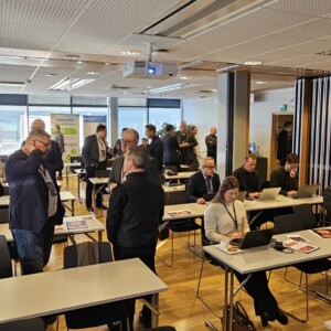 Joukko ihmisiä, enimmäkseen liikemiesten vaatteissa, seisoo ja istuu modernissa kokoushuoneessa, jossa on suuret ikkunat. Jotkut työskentelevät kannettavien tietokoneiden parissa pöydissä, kun taas toiset keskustelevat tai valmistautuvat kokoukseen.