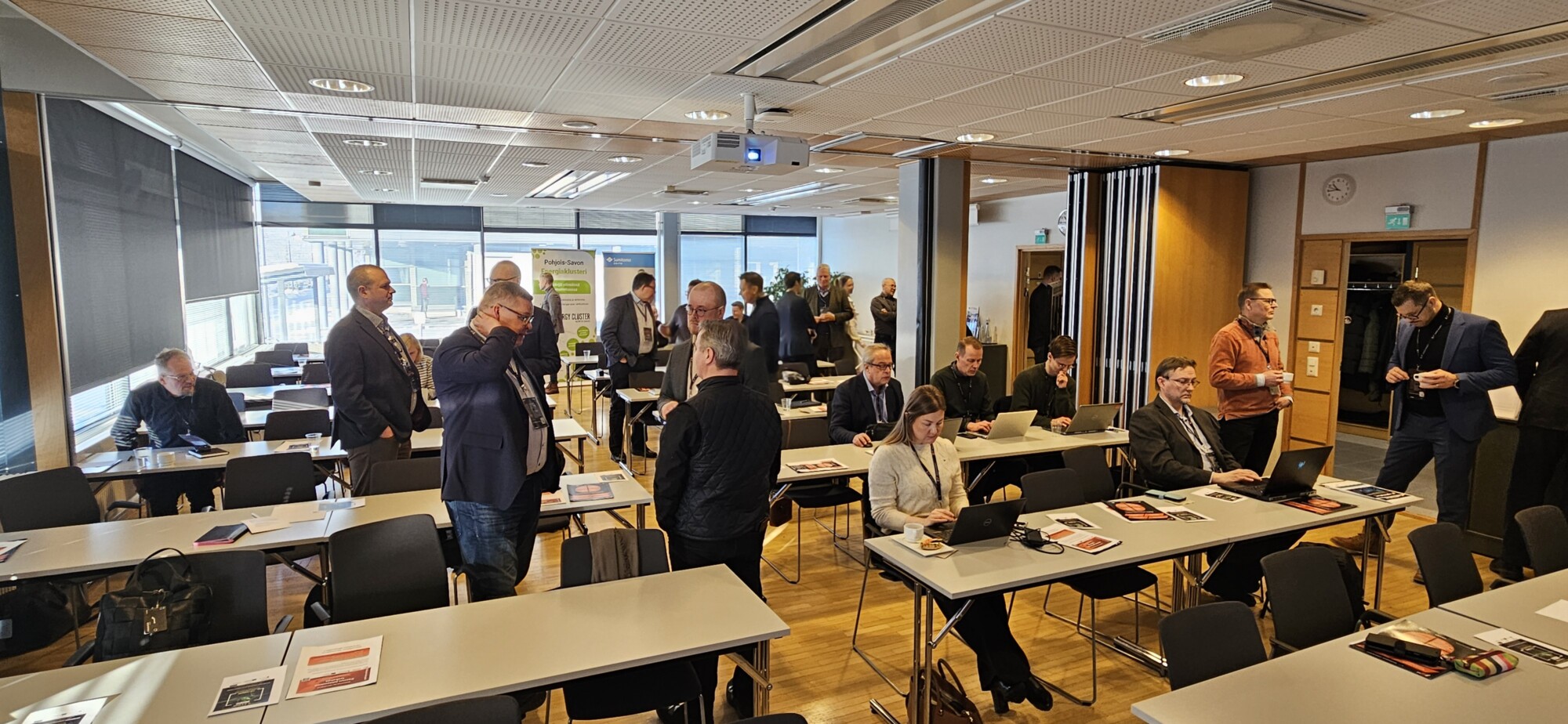 Joukko ihmisiä, enimmäkseen liikemiesten vaatteissa, seisoo ja istuu modernissa kokoushuoneessa, jossa on suuret ikkunat. Jotkut työskentelevät kannettavien tietokoneiden parissa pöydissä, kun taas toiset keskustelevat tai valmistautuvat kokoukseen.