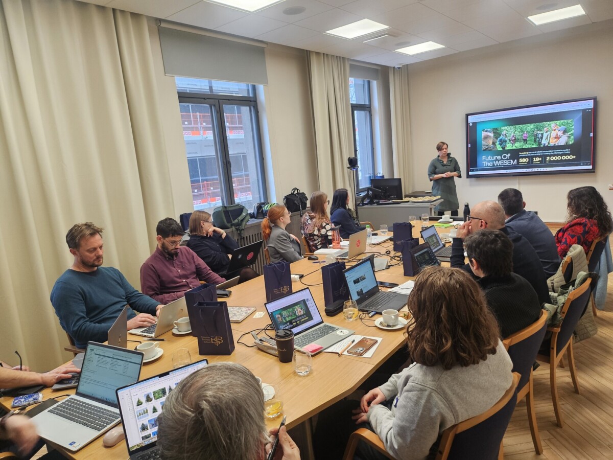 Joukko ihmisiä istuu neuvottelupöydän ympärillä kannettavien tietokoneiden ja muistikirjojen kanssa ja kuuntelee esitelmöitsijää, joka seisoo näytön vieressä, jossa näkyy Return of the Museum. Huoneessa on suuret ikkunat ja beiget verhot.