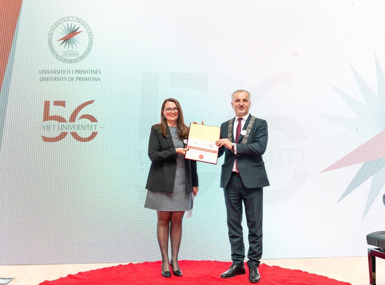 Virallisesti pukeutuneet nainen ja mies seisovat punaisella matolla ja pitelevät todistusta yhdessä taustan edessä, jossa on Prishtinan yliopiston logo ja 56 VJET UNIVERSITET -teksti.