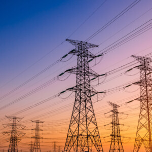 Korkeat sähkönsiirtomastot, joissa on useita voimalinjoja, ulottuvat kaukaisuuteen auringonlaskun värikkäällä taivaalla, jossa sekoittuvat sinisen, oranssin ja keltaisen sävyt.