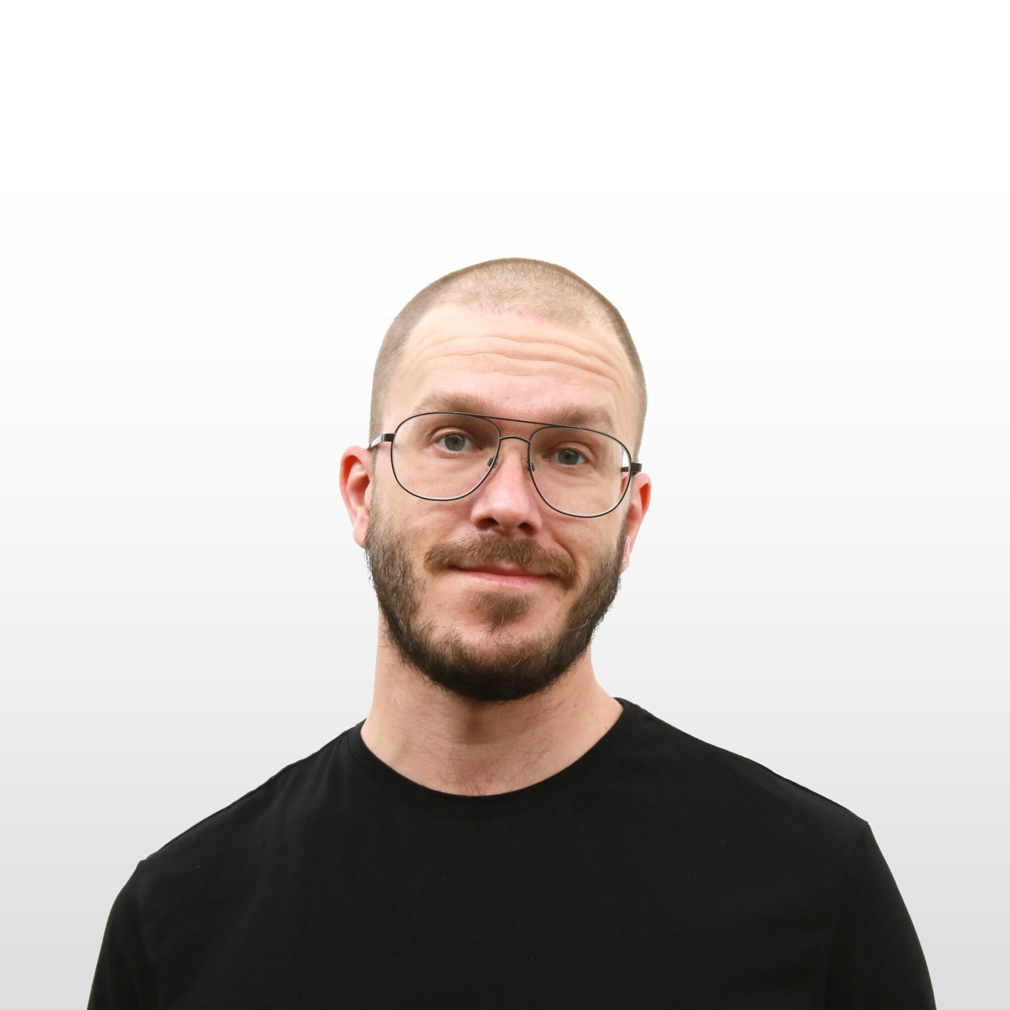 Mies, jolla on ajeltu pää, parta ja silmälasit sekä musta t-paita, seisoo vaaleanharmaata taustaa vasten ja katsoo kameraan neutraalilla ilmeellä.