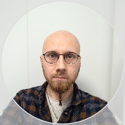 Kalju, täyspartainen ja silmälasipäinen henkilö, jolla on ruudullinen paita, katsoo suoraan kameraan valkoista taustaa vasten.