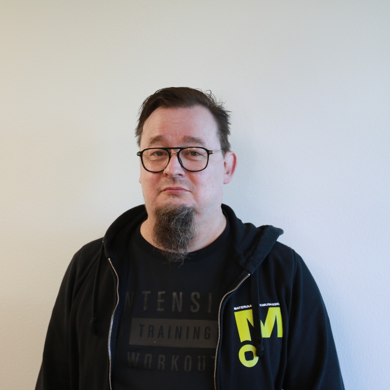 Silmälasipäinen mies, jolla on lyhyet tummat hiukset ja pukinparta, seisoo valkoista seinää vasten. Hänellä on yllään musta vetoketjullinen huppari mustan T-paidan päällä, jossa on keltaisia ja valkoisia kirjaimia.