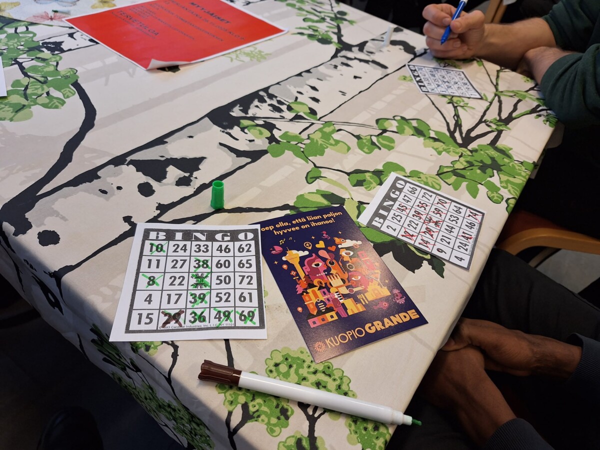 Kaksi ihmistä istuu pöydän ääressä, joka on peitetty lehtien peittämällä pöytäliinalla, ja pelaa bingoa. Pöydällä on bingokortteja, tusseja ja kyniä sekä värikäs kortti, jossa on kuvitus taloista.