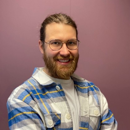 Hymyilevä henkilö, jolla on silmälasit ja parta, seisoo yksinkertaisen violetin taustan edessä, ja hänellä on yllään valkoinen t-paita ja sinikeltainen, keltainen ja harmaa ruutupaita.