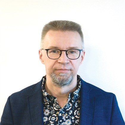 Keski-ikäinen mies, jolla on lyhyet, vaaleanruskeat hiukset, silmälasit ja leikattu harmaa parta, ja jolla on tummansininen bleiseri ja kukkapaita, seisoo valkoista taustaa vasten.