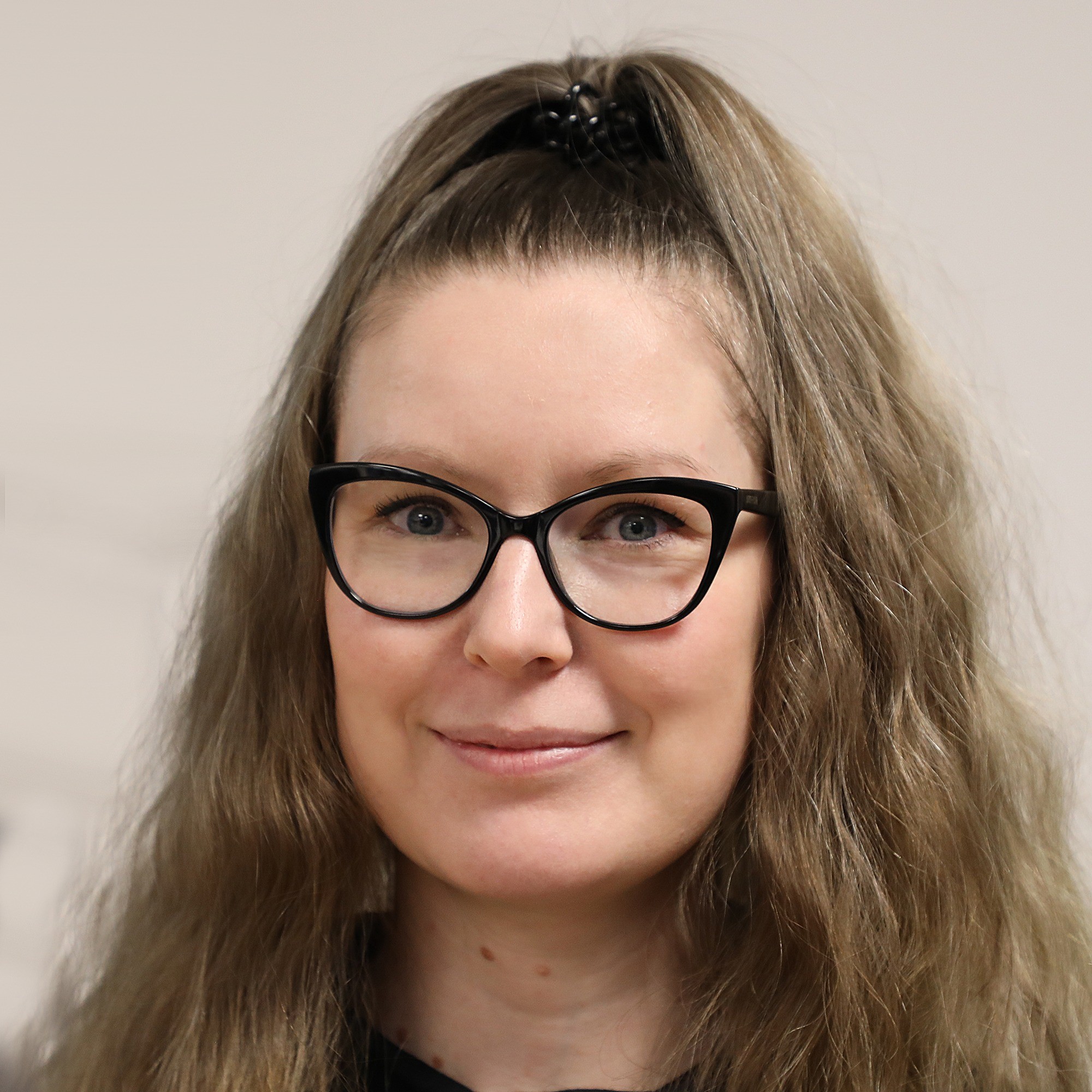 Nainen, jolla on pitkät, vaaleanruskeat, aaltoilevat hiukset, mustat silmälasit ja musta toppi, hymyilee kameralle. Hänen hiuksensa on vedetty taakse hiusklipsillä, ja tausta on tavallinen ja vaalea.