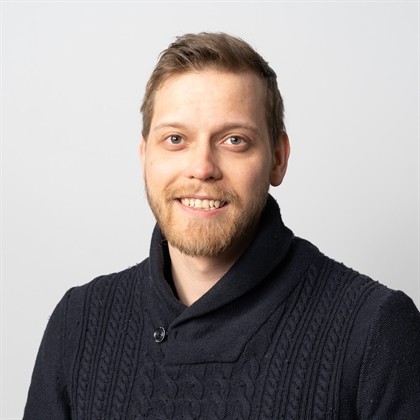 Hymyilevä mies, jolla on lyhyet vaaleanruskeat hiukset ja parta, ja jolla on yllään tumma pusero, poseeraa vaaleanharmaan taustan edessä.