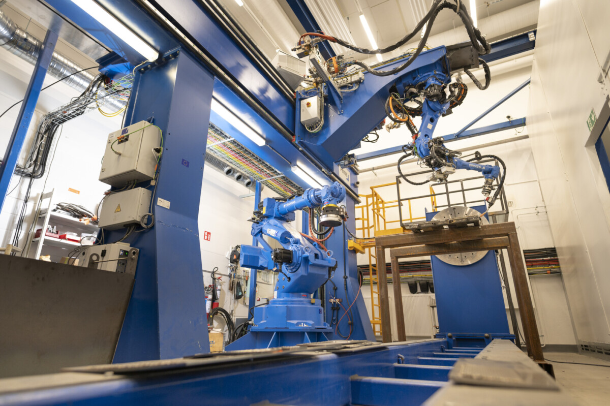 Teollisuusrobotit tehdasympäristössä, jossa siniset robottikäsivarret työskentelevät metallirakenteessa, jota ympäröivät kaapelit ja koneet kirkkaiden kattovalojen alla.