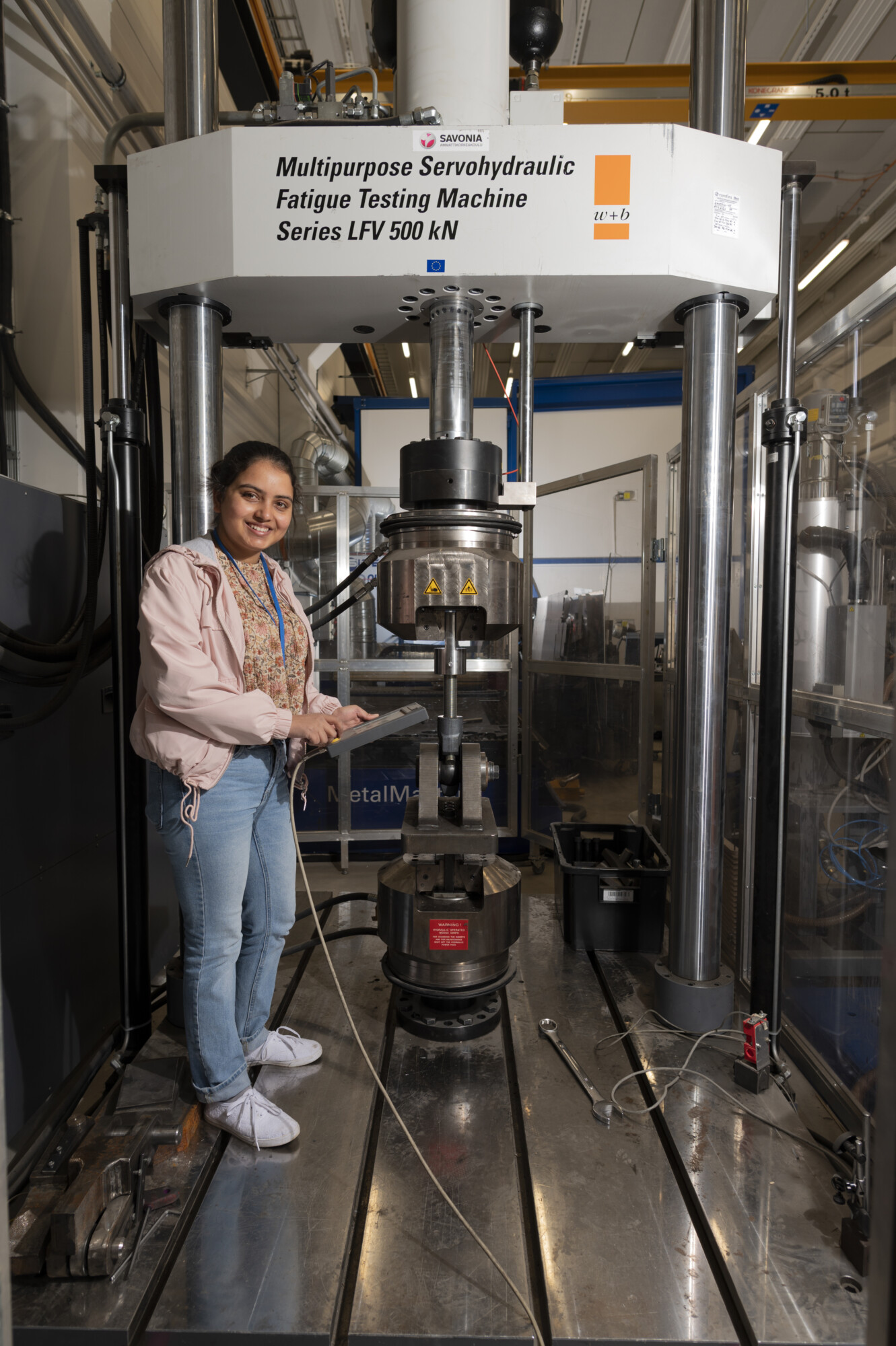Nainen seisoo laboratoriossa suuren monikäyttöisen servohydraulisen väsymistestauskoneen vieressä hymyillen ja elehtien kohti laitetta.