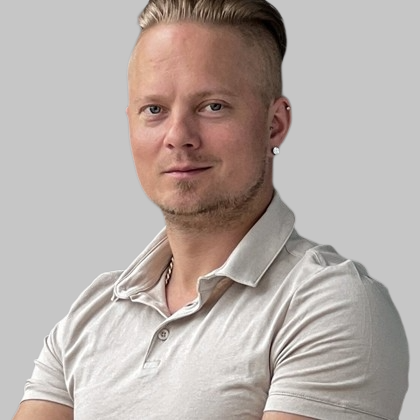 Lyhyet, vaaleat hiukset ja vaaleat kasvohiukset omaavalla miehellä on yllään vaalean beige poolopaita. Hän seisoo kädet ristissä ja katsoo kameraan vaaleanharmaata taustaa vasten.