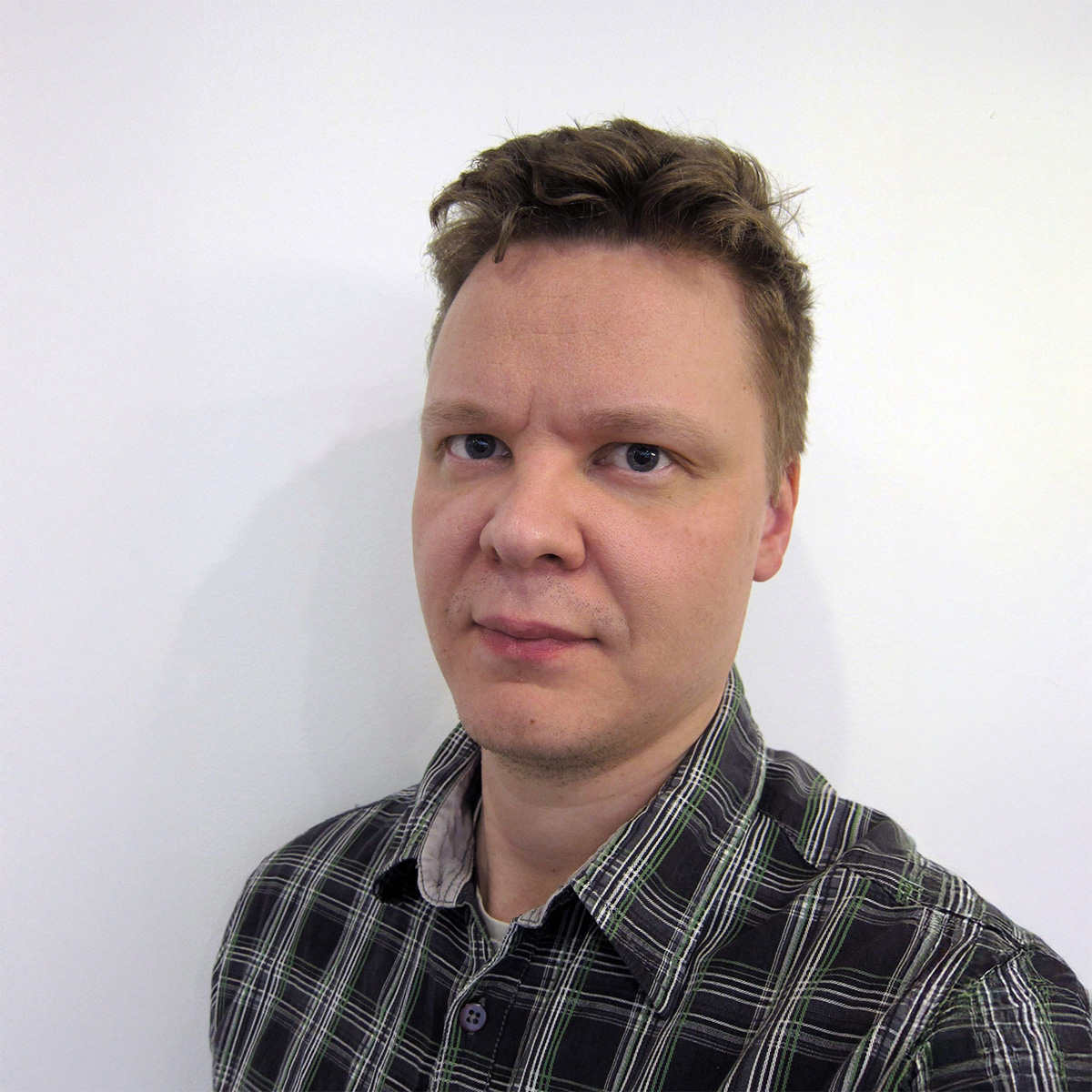 Lyhyet, vaaleanruskeat hiukset omaava henkilö seisoo valkoisen taustan edessä, yllään ruudullinen paita ja katsoo kameraan neutraalilla ilmeellä.