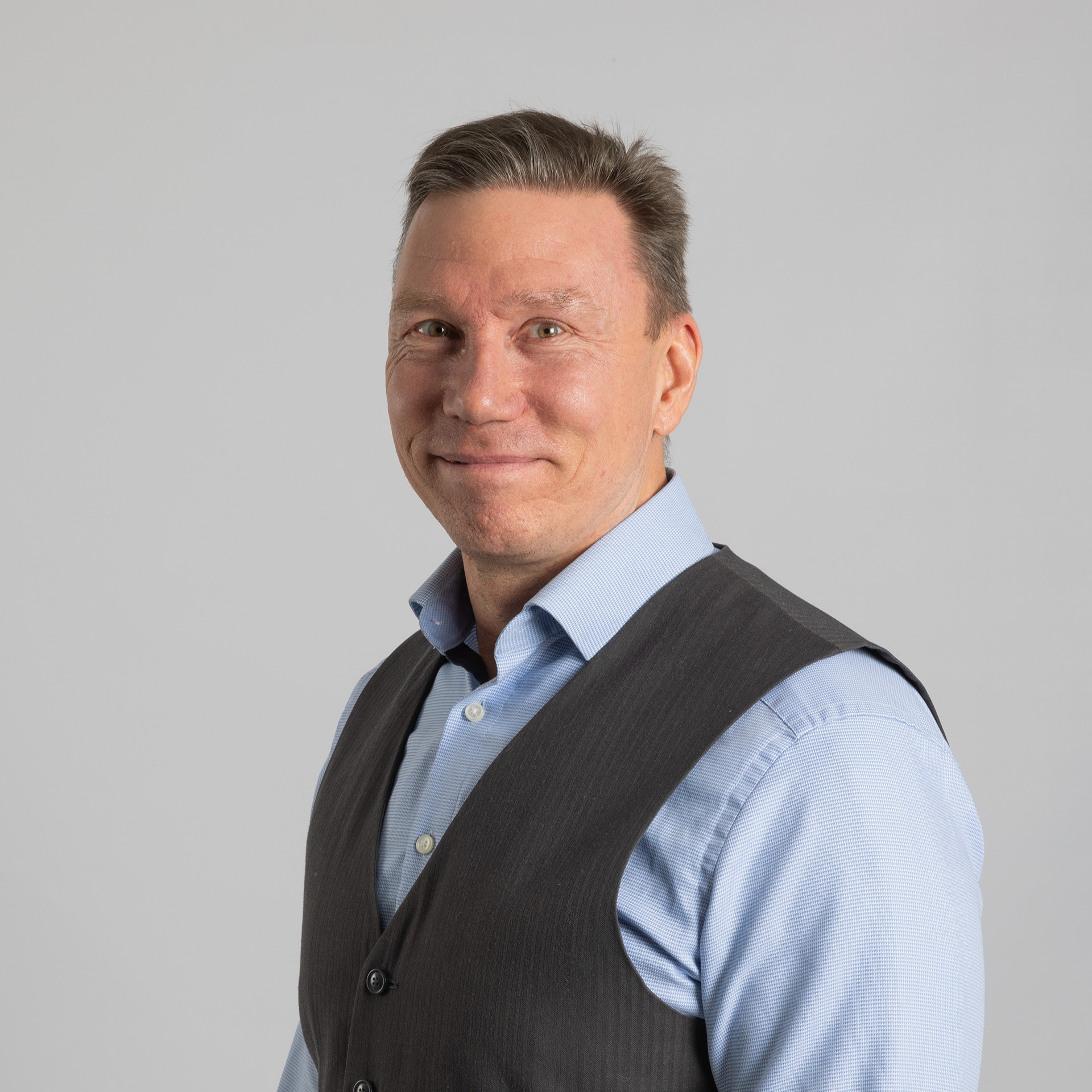 Lyhyet ruskeat hiukset omaava mies hymyilee ja katsoo kameraan. Hänellä on vaaleansininen paita ja tummanharmaa liivi, ja hän seisoo vaaleanharmaata taustaa vasten.