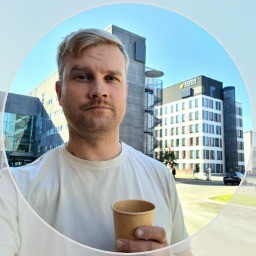 Lyhyet vaaleat hiukset ja viikset omaava mies, jolla on valkoinen t-paita, pitelee paperimukia seisoessaan ulkona modernin toimistorakennuksen edessä aurinkoisena päivänä.
