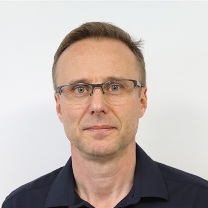 Keski-ikäinen mies, jolla on lyhyet vaaleanruskeat hiukset, suorakulmaiset silmälasit ja tumma kauluspaita, seisoo valkoisen taustan edessä.