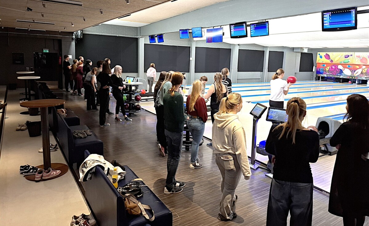 Joukko ihmisiä, enimmäkseen naisia, seisoo ja katsoo, kun muut keilaavat kirkkaasti valaistulla keilaradalla. Jotkut valmistautuvat keilaamaan, kun taas toiset seurustelevat penkkien lähellä, ja kengät ovat lattialla lähellä.