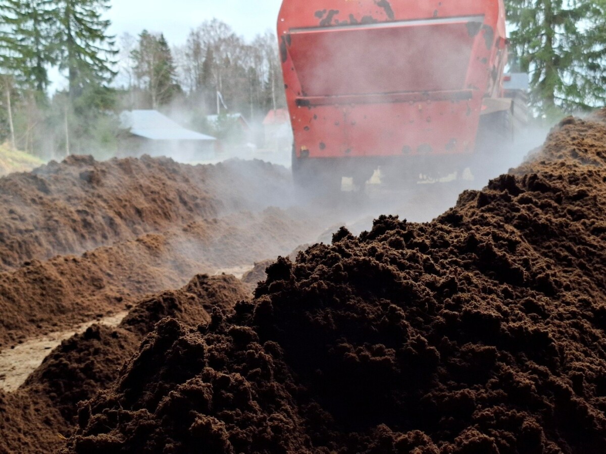 Lähikuva höyryävästä maasta, jota siirretään tai levitetään punaisella maatalouskoneella ulkona maaseudulla, taustalla puita ja rakennuksia.