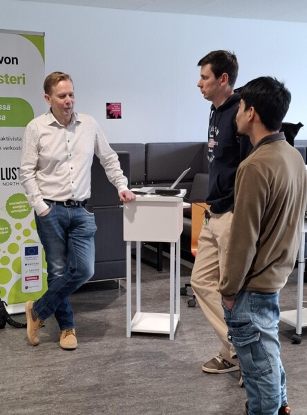 Kolme miestä seisoo keskustelemassa pienen valkoisen korokkeen vieressä sisätiloissa. Yksi mies nojaa koroketta vasten, kun kaksi muuta kääntyvät häntä kohti, ja taustalla näkyy pystysuora vihreä-valkoinen banderolli.