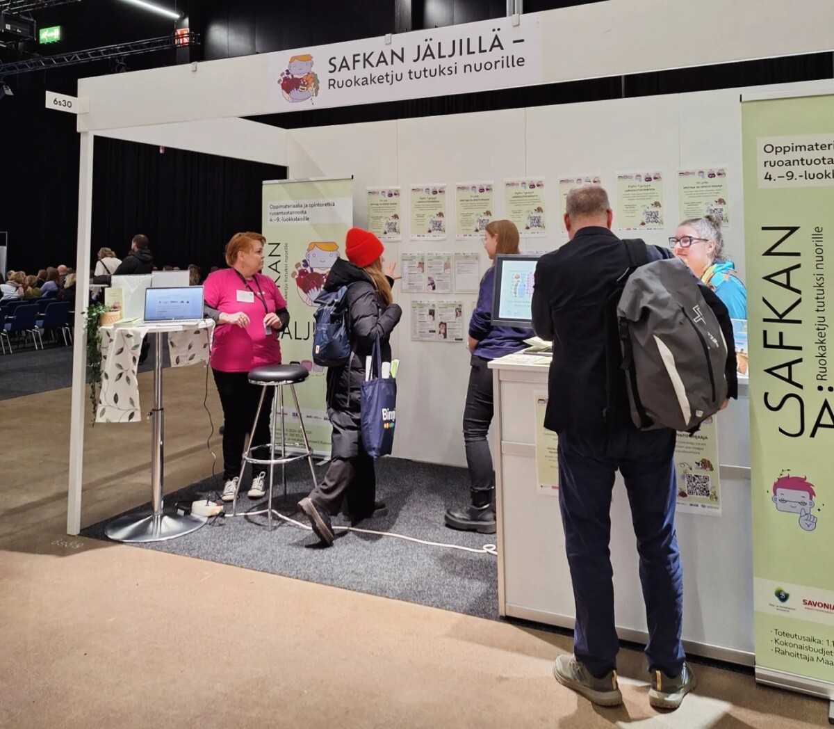 Ihmisiä vuorovaikutuksessa näyttelyosastolla, jossa on kyltti SAFKAN JÄLJILLÄ - Ruokaketju tutuksi nuorille. Esillä on julisteita ja monitori, ja kaksi henkilökunnan jäsentä keskustelee kävijöiden kanssa.