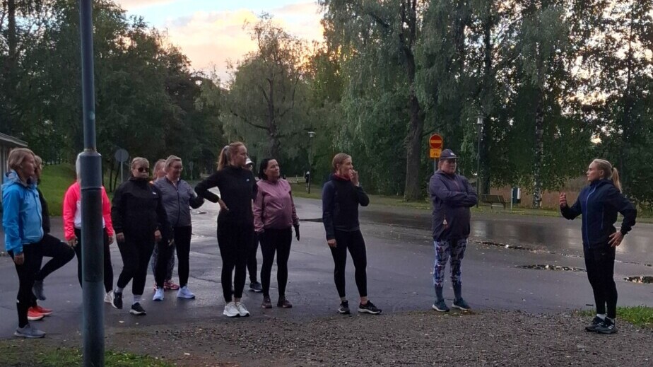 Joukko urheiluvaatteisiin pukeutuneita ihmisiä seisoo ja keskustelee kadun risteyksen lähellä ulkona, taustalla puita ja pilvinen taivas.