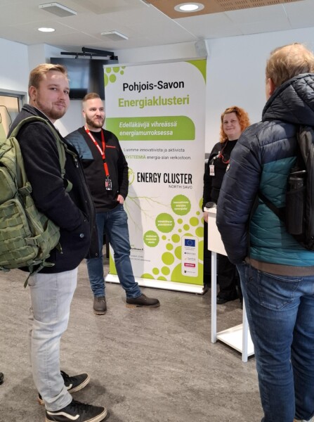 Neljä ihmistä seisoo ja keskustelee kojun vieressä, jossa lukee Pohjois-Savon Energiaklusteri ja Energiaklusteri Pohjois-Savo. Koju on sisätiloissa, ja kahdella henkilöllä on tiskin takana mustat paidat ja kaulanauhat.