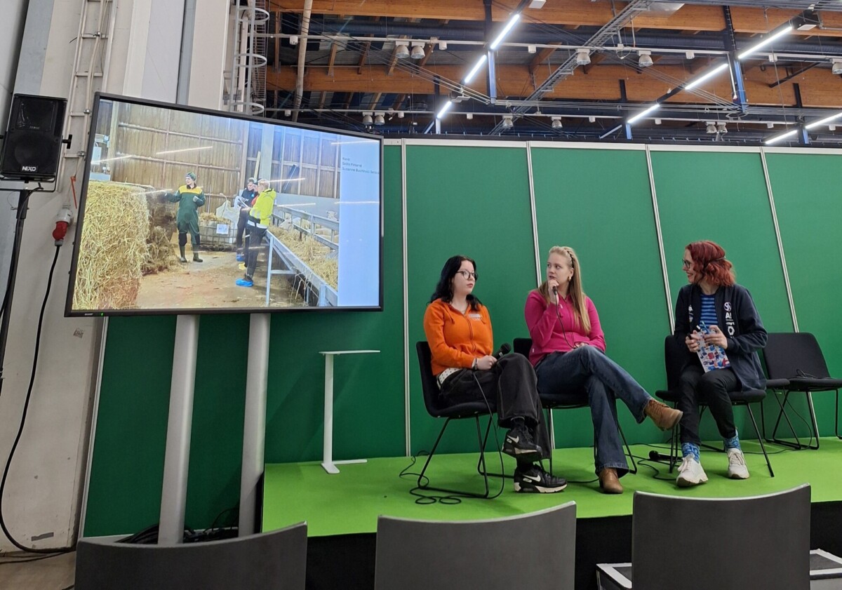 Kolme ihmistä istuu lavalla keskustelemassa. Heidän takanaan näytöllä näkyy kuva kahdesta ihmisestä, jotka käsittelevät heinää ladossa. Taustalla on vihreitä paneeleita ja yläpuolella teollisuusvalaistus.