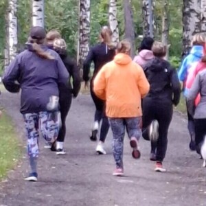 Joukko urheiluvaatteisiin pukeutuneita ihmisiä hölkkäilee puistossa puiden reunustamaa polkua pitkin, jota ympäröivät korkeat koivut, joiden lehdet ovat vihreitä. Kohtaus on ulkona ja sää näyttää viileältä.