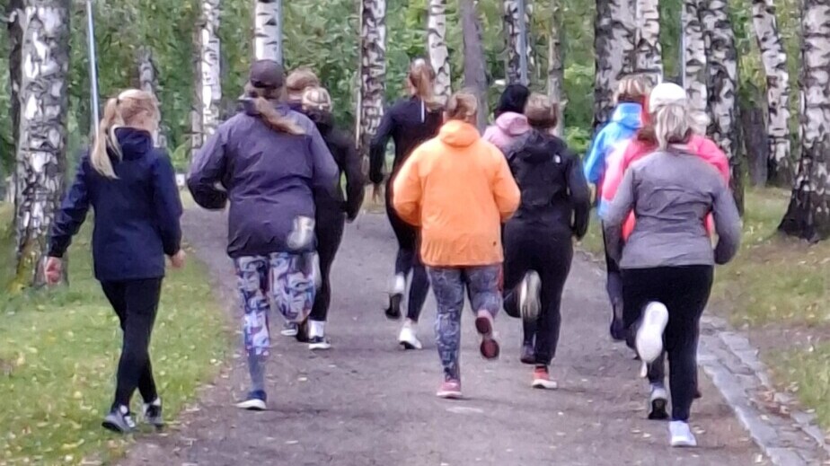 Joukko urheiluvaatteisiin pukeutuneita ihmisiä hölkkäilee puistossa puiden reunustamaa polkua pitkin, jota ympäröivät korkeat koivut, joiden lehdet ovat vihreitä. Kohtaus on ulkona ja sää näyttää viileältä.